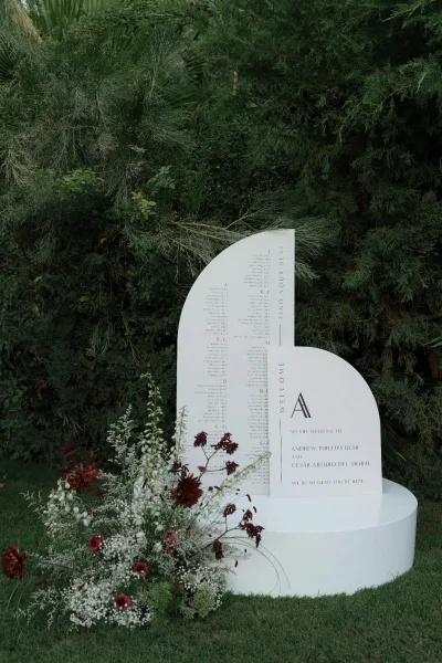Wedding welcome sign with curved white panel and seating chart text, framed by baby's breath and red flowers in a garden setting