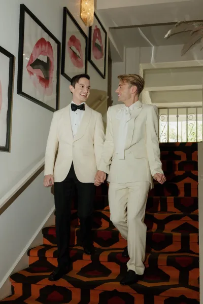 Couple portrait of two grooms holding hands in cream suits, descending a staircase with patterned runner and chandelier overhead