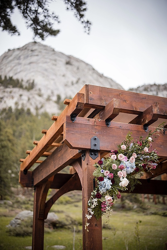 Wedding ceremony arch with a wooden wedding arch draped in roses, hydrangea and greenery, set in a mountain meadow with pines and sky