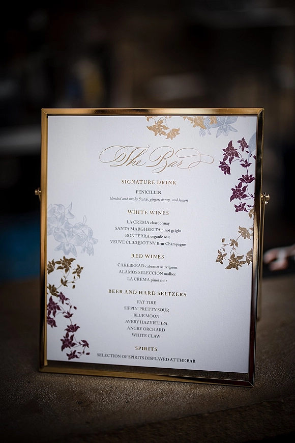 Wedding bar menu and signature drink sign in a gold frame with floral illustration and calligraphy lettering on a bar counter indoors