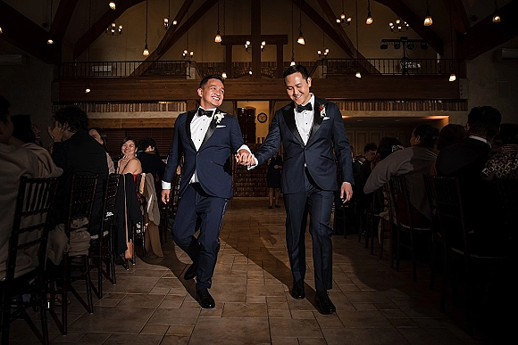 Wedding recessional as a same sex wedding couple of two grooms hold hands in tuxedos, walking past guests in a pendant-lit hall
