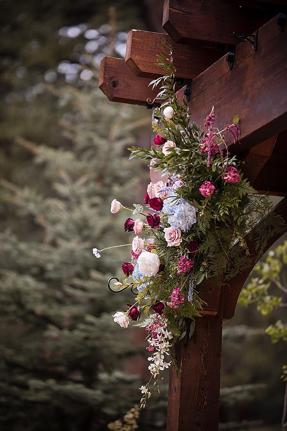 Wedding arbor flowers spill in an asymmetrical floral arbor arrangement of roses and hydrangea on a wooden arch, set by evergreen trees
