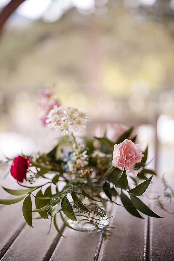 Wedding bouquet in a glass vase with pink carnation and red ranunculus, white blooms and greenery on a wood table with bokeh lights