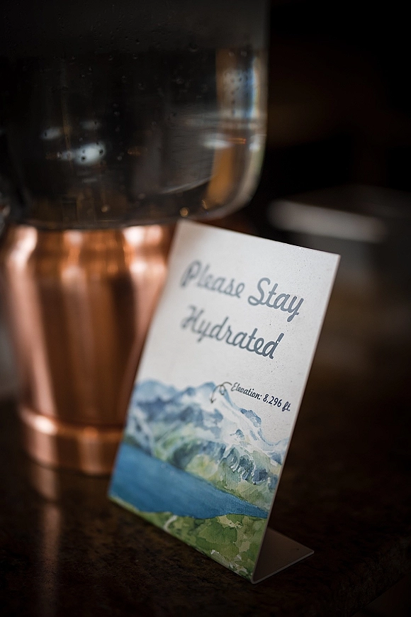 Wedding hydration sign with watercolor illustration on a tent card beside a copper beverage urn at an indoor reception countertop