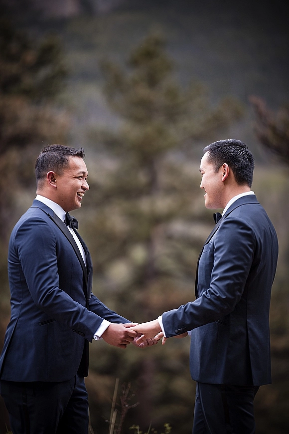 Wedding first look as two grooms in tuxedos hold hands and face each other, smiling in a softly blurred forest setting