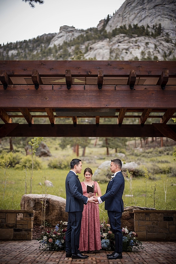 Wedding ceremony with officiant and grooms holding hands in navy suits beneath a wooden pergola, with stone pillars and mountain meadow backdrop