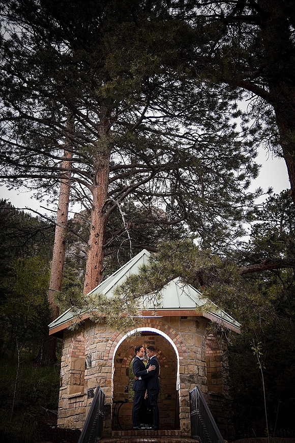 Couple portrait of two grooms in dark suits with boutonnieres, standing under a stone archway by a bridge railing in a forested mountain setting