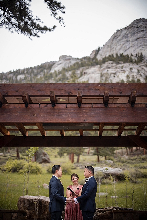 Wedding vows as two grooms hold hands while a bridesmaid officiant reads from a vow book under a wood pergola in the mountains