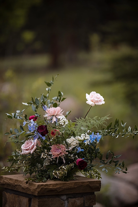 Wedding floral arrangement with roses and blue flowers spilling over a stone pedestal, framed by greenery in a garden with trees and lawn
