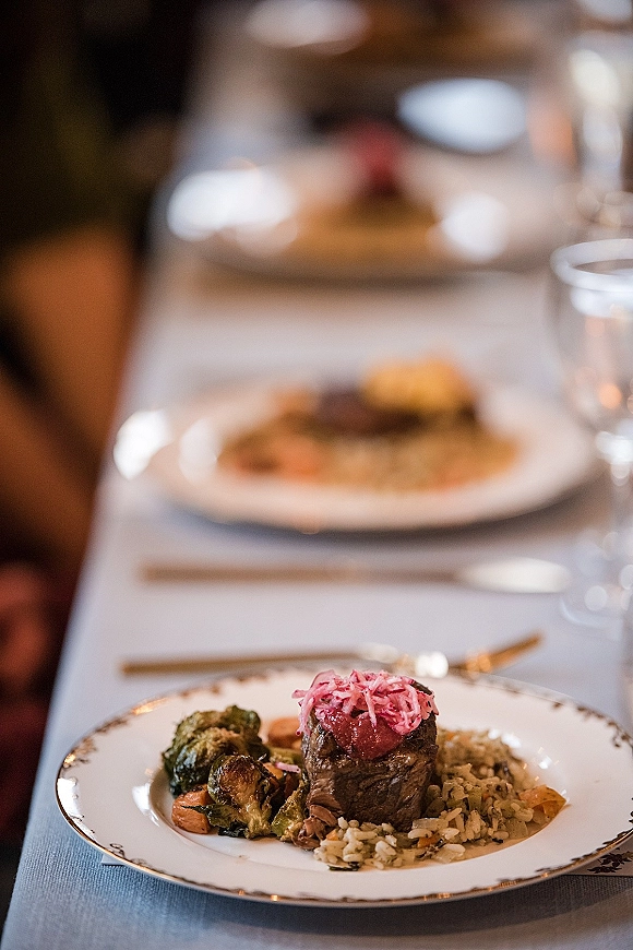 Wedding reception dinner with a plated wedding dinner entree on a gold-rimmed plate, silverware and water glass on a long banquet table