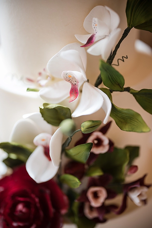 Orchid bouquet with white orchids and green leaves, accented by burgundy blooms, arranged against a neutral wall backdrop