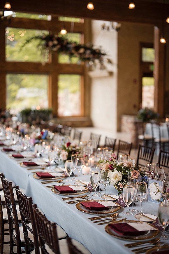 Reception tablescape with long banquet wedding table in light blue linens, burgundy napkins, gold rim plates, candles, florals, and pendant lights by large windows
