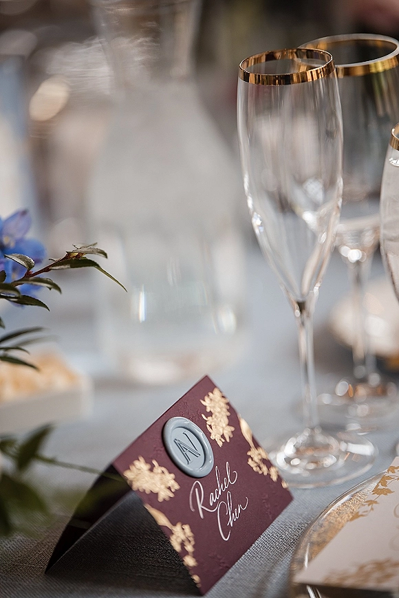 Wedding place card with wax seal and gold foil lettering beside a gold-rim champagne flute on a reception table with greenery accents