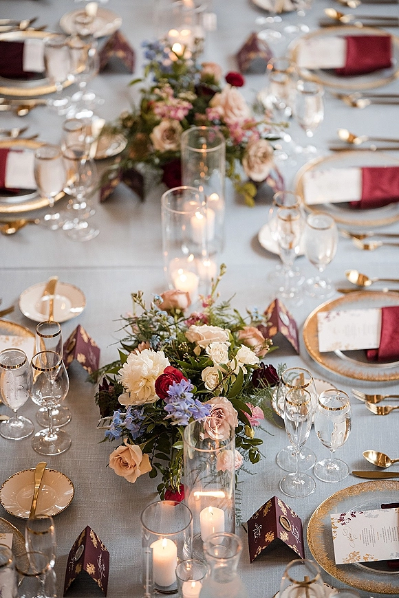 Reception tablescape with wedding table centerpiece of low florals and pillar candles in glass vases, gold-rim chargers, burgundy napkins on white cloth