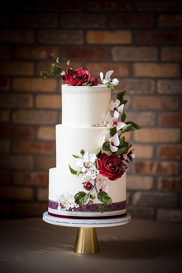 Wedding cake with three tier wedding cake design in white frosting, adorned with red roses, white orchids, and greenery on a stand against brick wall