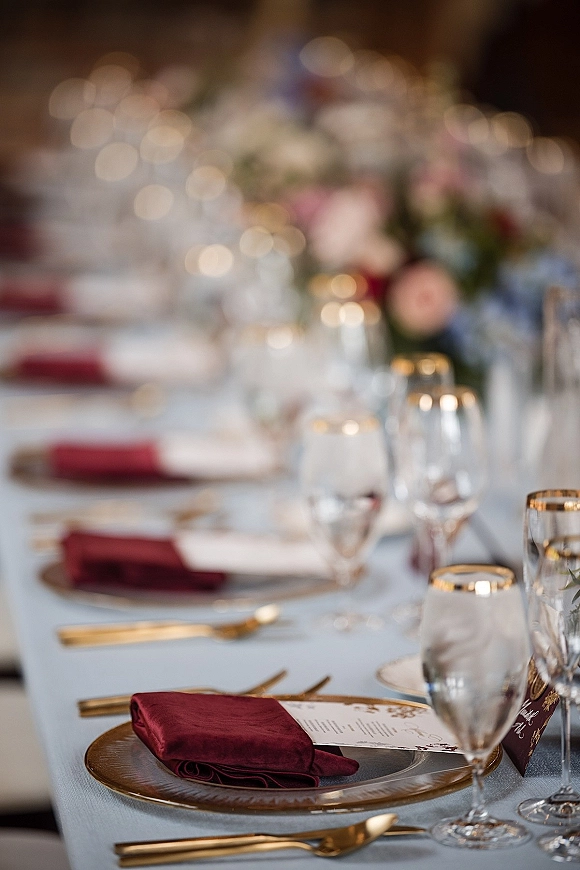 Reception tablescape with burgundy wedding tablescape details, gold flatware, amber charger plates, menus and place cards on a long banquet table with bokeh lights