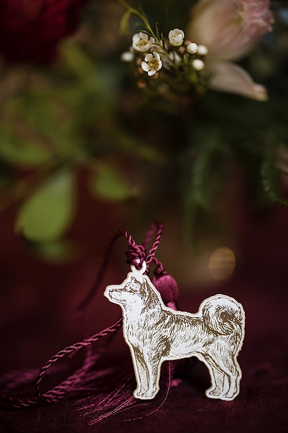 Wedding favor tag with a dog-shaped paper tag, purple cord, and tassel resting on burgundy fabric with blurred flowers and greenery behind