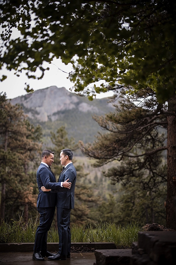 Couple portrait of two grooms holding each other in matching suits with bow ties, framed by pine trees and a mountain view under clouds