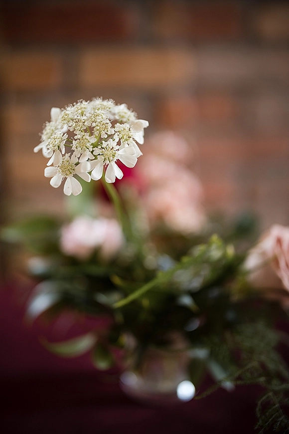 Wedding centerpiece with wildflower wedding centerpiece blooms in a glass vase, featuring a white lace flower on a burgundy tablecloth by a brick wall