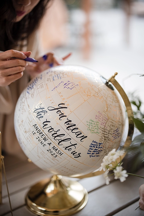 Wedding guest book globe on a gold stand with marker pen and handwritten messages on a table with greenery, white flowers, blurred guests behind