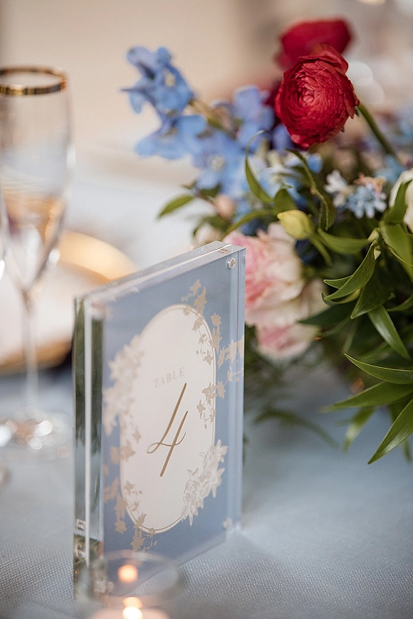 Wedding table number in an acrylic table number frame beside red ranunculus, blue delphinium, greenery, votive candles, and champagne flute