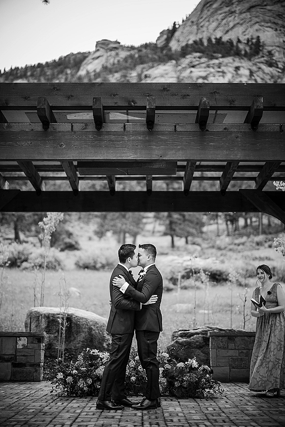 Wedding kiss between two grooms in matching suits with boutonnieres beneath a wooden pergola on brick pavers, mountains beyond