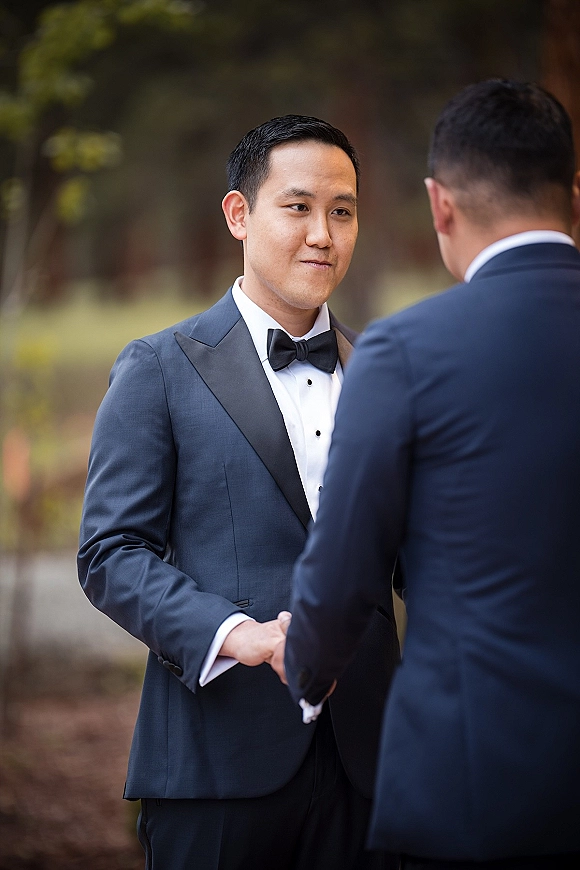 Groom portrait in a tuxedo with black bow tie, crisp dress shirt and cufflinks, standing outdoors against leafy green trees