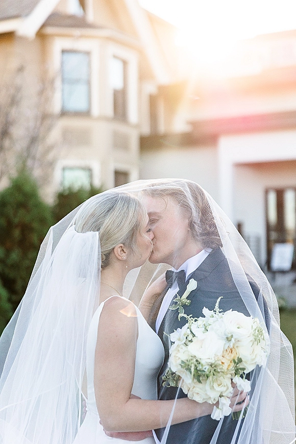 Wedding kiss portrait of bride and groom kissing under a veil, bride holding a white bouquet, sunlight by a house exterior