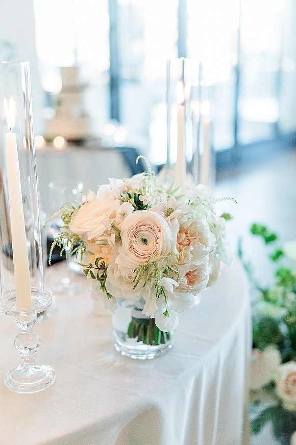 Wedding centerpiece with white floral centerpiece blooms, greenery, glass cylinder vases and taper candles on a white tablecloth by a cake table