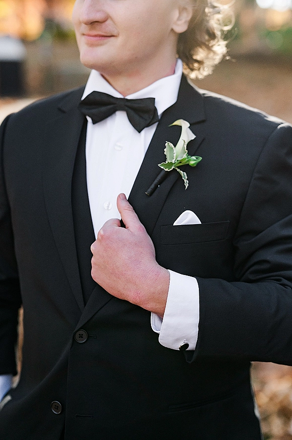 Groom portrait in a black tuxedo groom look, adjusting his jacket with bow tie and boutonniere against blurred outdoor foliage backdrop
