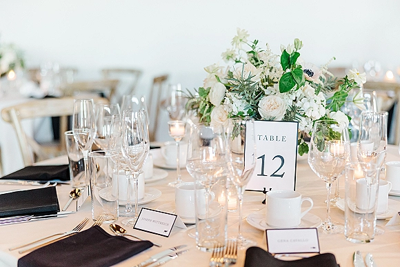 Reception tablescape with a wedding table centerpiece of white flowers and greenery, gold flatware, black napkins, and votive candles on white linens