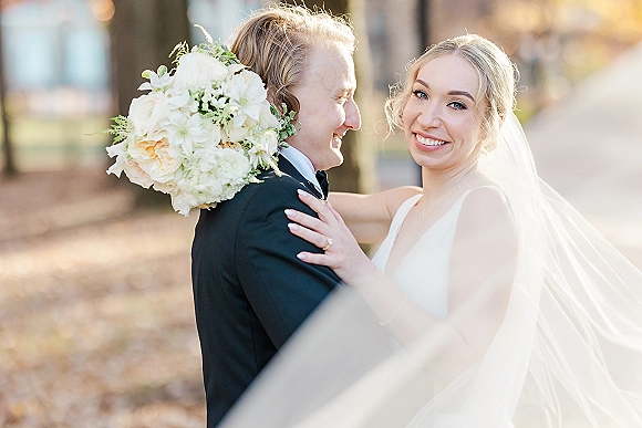 Couple portrait of bride and groom embrace, bride smiling at camera with bouquet and veil in golden autumn trees at sunset