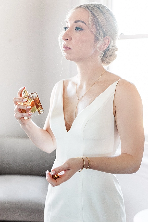 Bridal portrait of a bride getting ready, spraying perfume in a sleek satin V-neck dress by a window-lit sofa and neutral wall