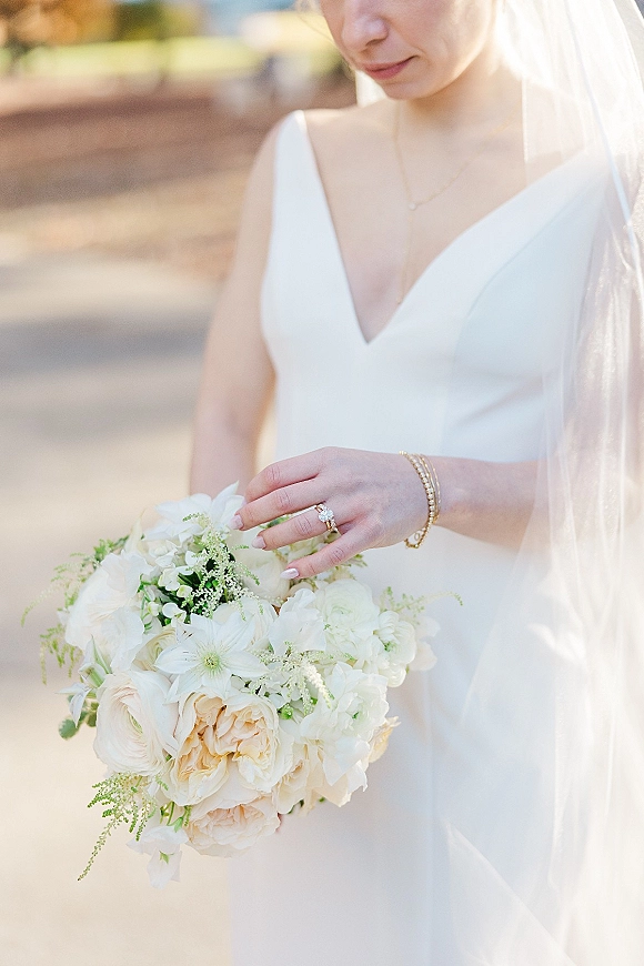 Bridal portrait of a bride holding bouquet of white and blush blooms, wearing a veil and white dress on an outdoor walkway with greenery