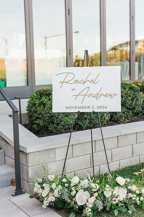 Wedding welcome sign on a black easel, acrylic with gold lettering, framed by white roses and anthurium at the base by glass doors