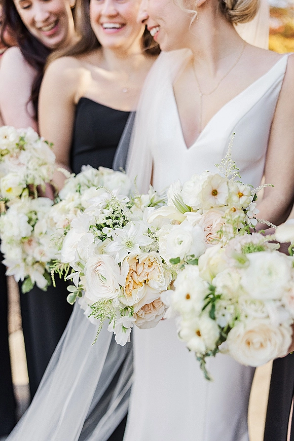 Bridesmaid portrait with bridesmaids holding bouquets of white and blush flowers with greenery beside the bride in veil, soft outdoor foliage backdrop