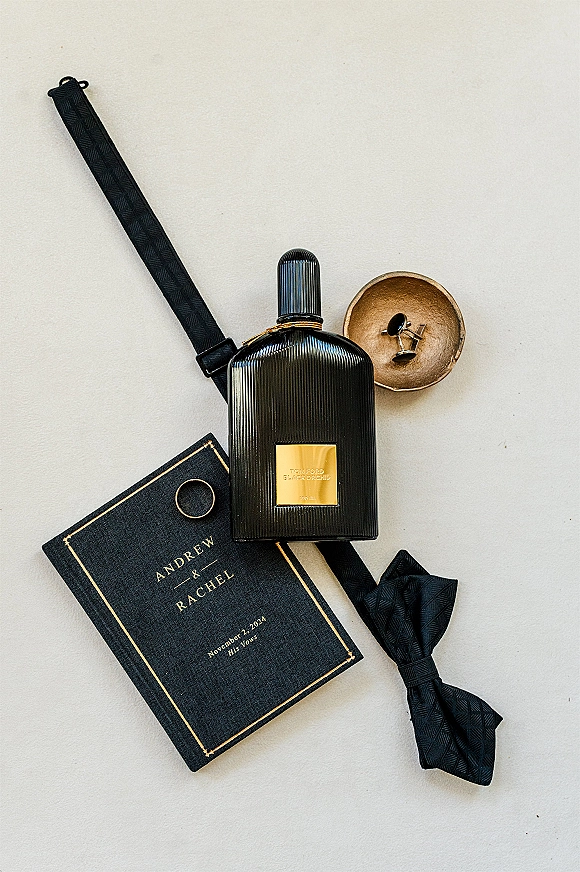 Groom details flatlay with cologne bottle, black bow tie, suspenders, cufflinks, wedding band and invitation arranged on a white surface