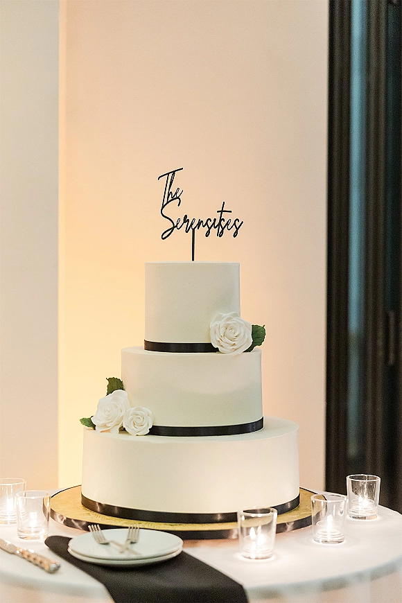 Wedding cake with black ribbon on a cake stand, topped with script topper, white sugar flowers and greenery, votive candles nearby