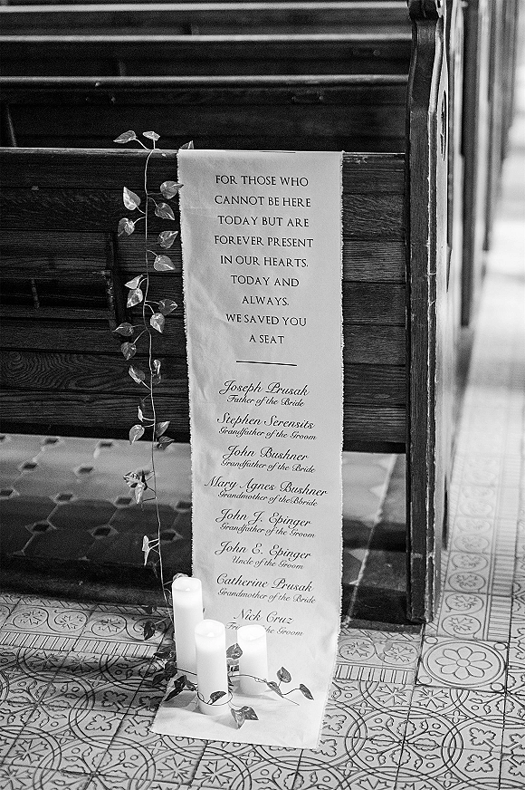 Wedding memorial sign with reserved seat wedding sign text, draped fabric and greenery vine beside pillar candles by wooden pews on tile floor