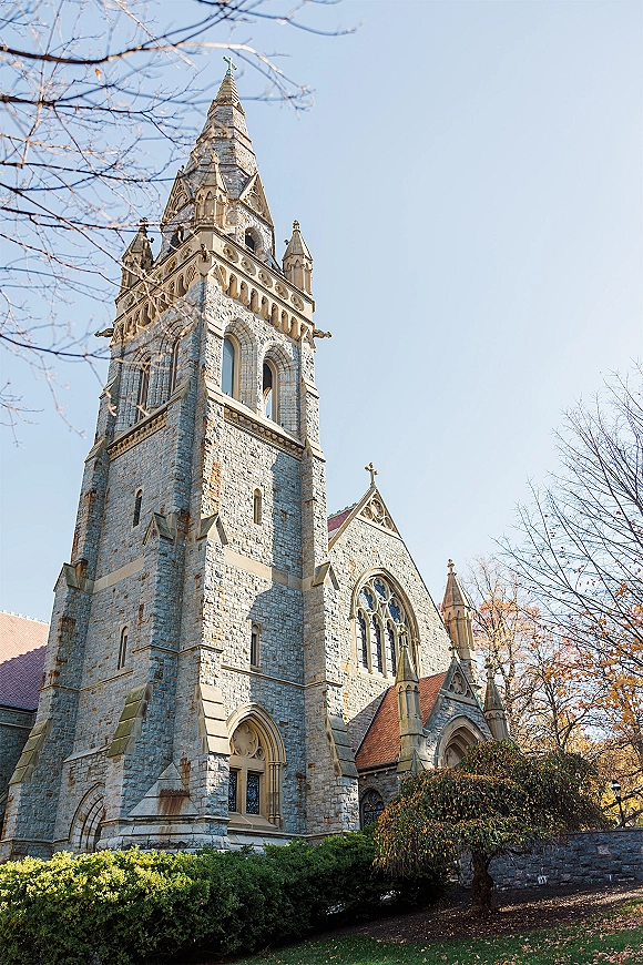 Church exterior with stone facade and steeple, arched stained glass windows and red roof tiles beneath a blue sky and autumn trees