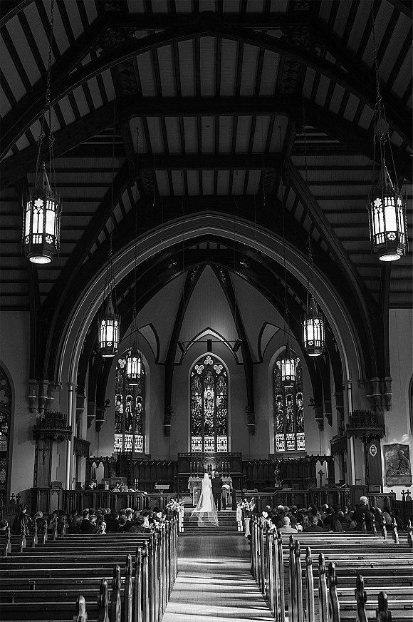 Ceremony moment at a church wedding ceremony, bride and groom at the altar seen from behind, with long veil and stained glass windows.