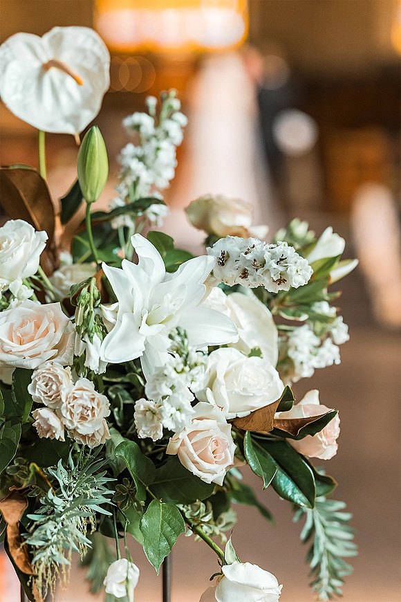 Wedding floral arrangement centerpiece of white lilies and blush roses with greenery, glowing under warm ambient lights in a blurred reception venue