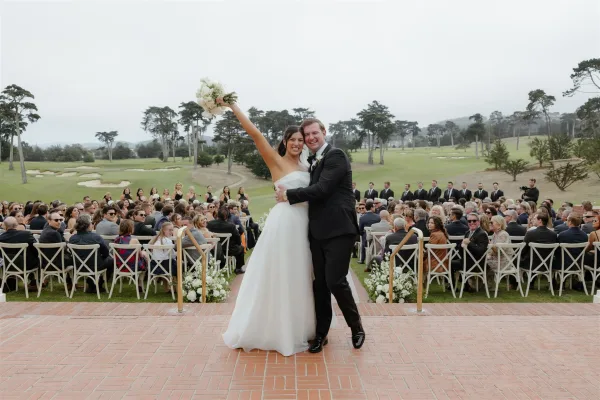 Ceremony recessional as newlyweds walk up the aisle, bride raising her bouquet, guests cheering beside white chairs on a golf course lawn