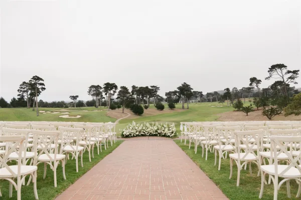 Ceremony aisle decor with white crossback chairs lining a brick aisle, ending at a low white-and-green floral arrangement on a golf course lawn