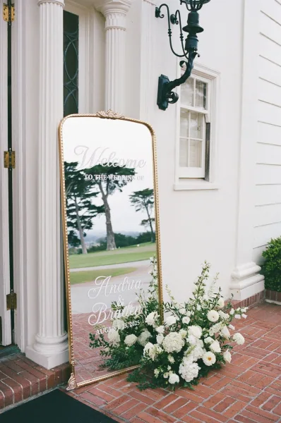 Wedding welcome sign on a gold ornate mirror with vinyl calligraphy and white roses, set on brick steps by a columned entrance