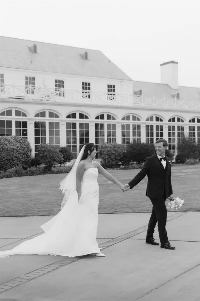 Couple portrait of bride and groom holding hands, bride in strapless dress with long veil and bouquet on a lawn by arched windows