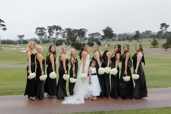 Bridal party portrait of the bride with bridesmaids in black dresses holding white rose bouquets on a golf course lawn under an overcast sky