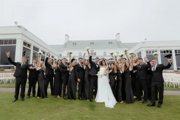 Wedding party portrait with bride holding a white bouquet, bridesmaids and groomsmen cheering on a lawn by a white building under overcast sky