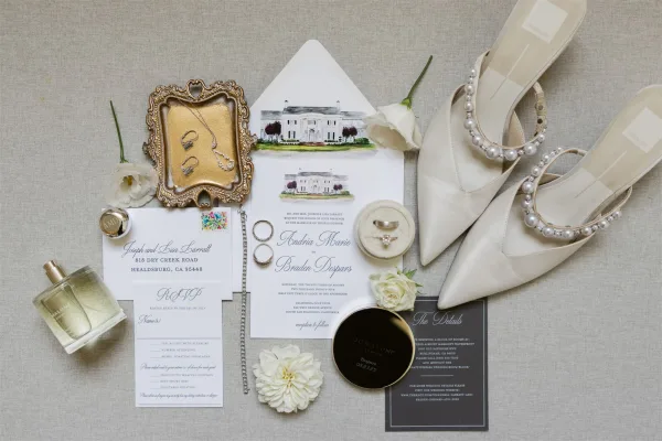 Invitation suite with watercolor venue invitation on a linen backdrop, styled with rings, pearl-strap bridal heels, perfume, wax seal and flowers