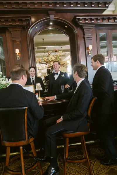 Groomsmen portrait in black tuxedos and bow ties, laughing on bar stools with champagne flute at a wood-paneled bar backdrop